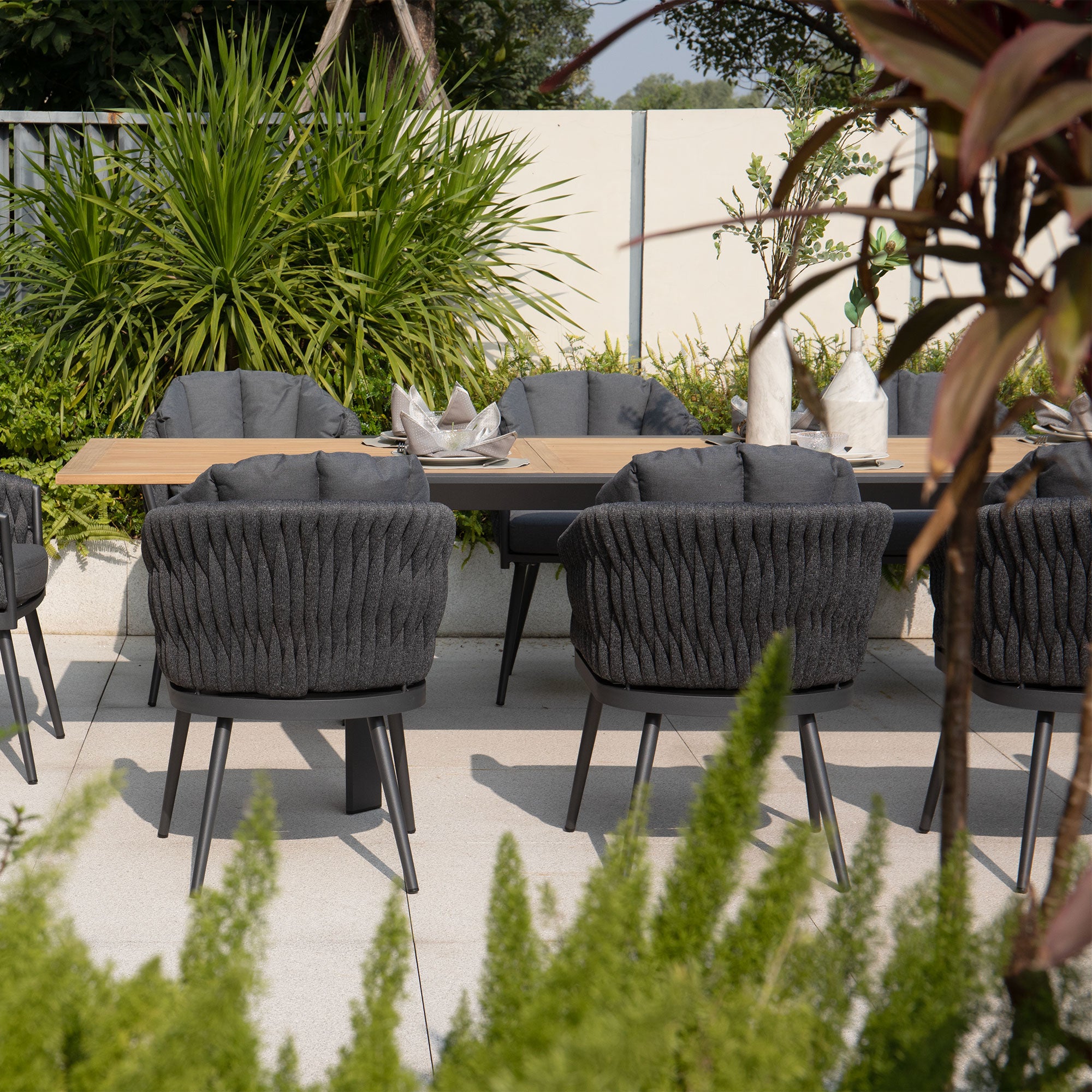 Outdoor dining area featuring a teak wooden table, ten black cushioned chairs, and lush green plants in the background and foreground. Perfect for any occasion, this Palma 10 Seat Rope Extending Teak Dining Set in Grey combines elegance with functionality.