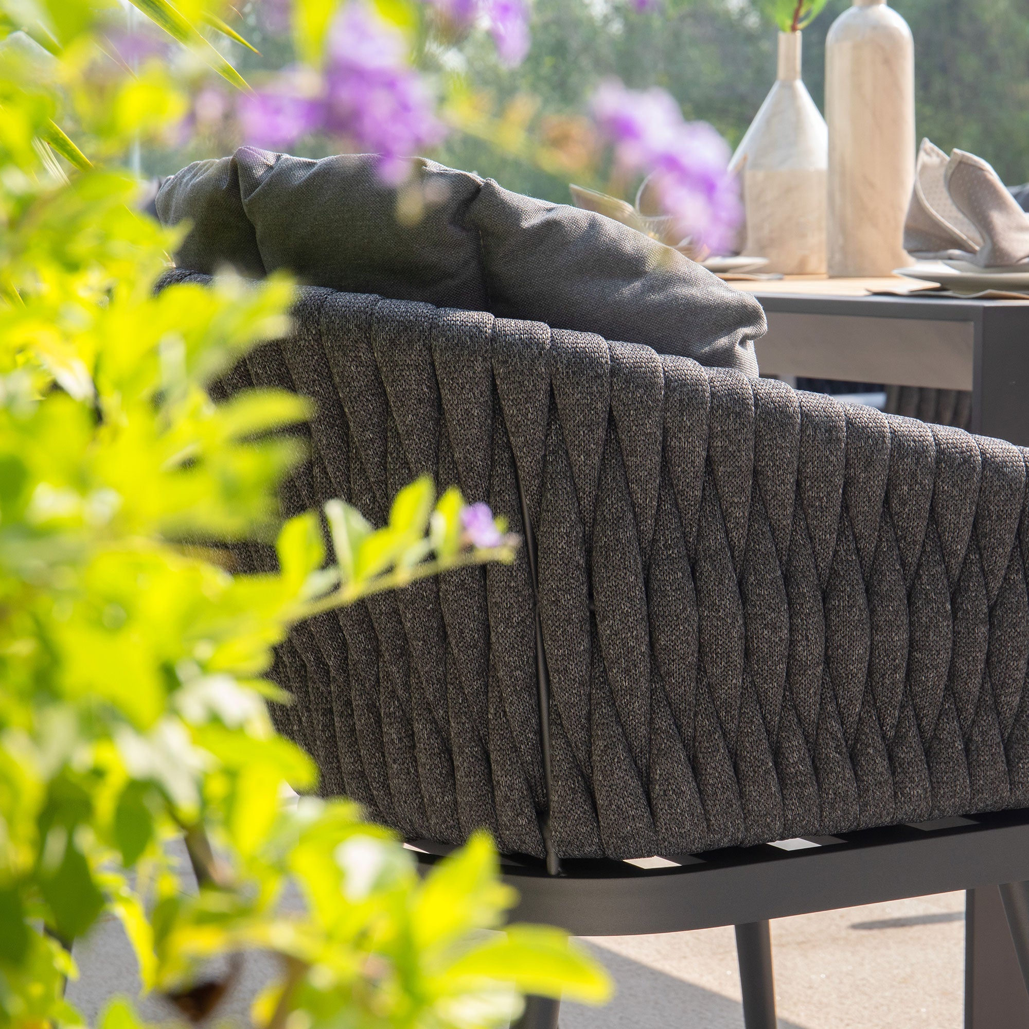 A cozy gray woven chair from the Harbour Lifestyle Palma collection sits on a patio, surrounded by vibrant greenery and purple flowers, perfectly complementing your Palma 10 Seat Rope Extending Teak Dining Set in Grey.