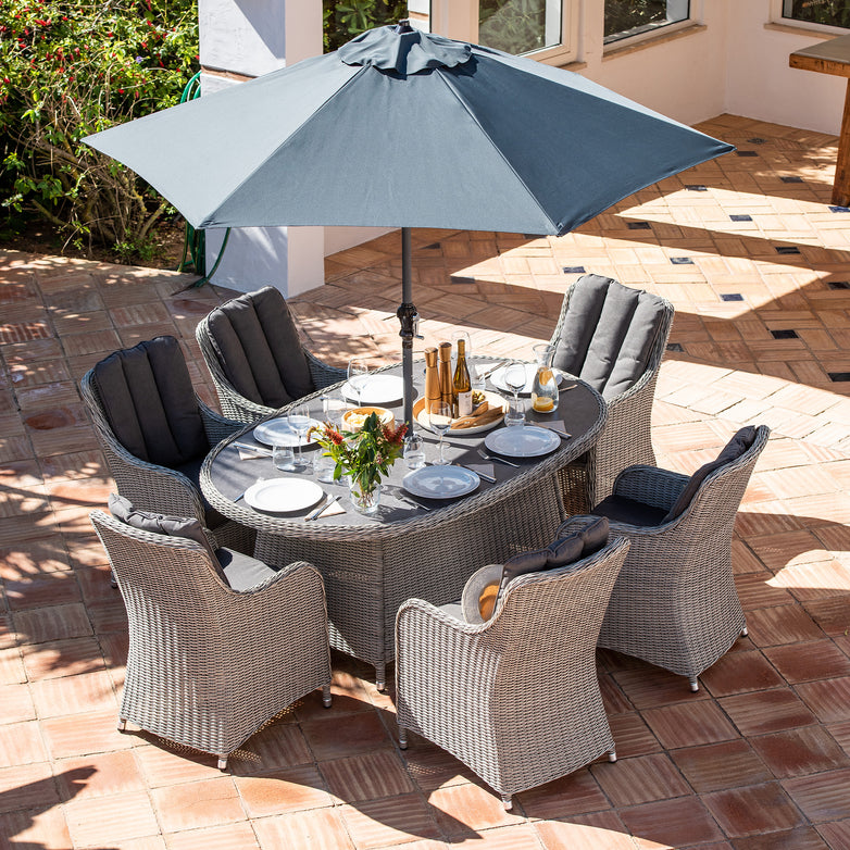 The Santiago 6 Seat Rattan Oval Dining Set in Grey features an outdoor patio dining set with a glass top table, aluminium powder-coated frames, six wicker chairs, and a large grey umbrella, making it perfect for a meal.