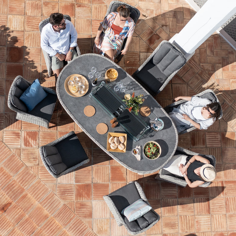 Four people sit around a Santiago 8 Seat Rattan Oval Firepit Dining Set in Grey with food and drinks, enjoying their time on a tiled patio. The set features durable aluminium powder-coated frames for long-lasting outdoor enjoyment.