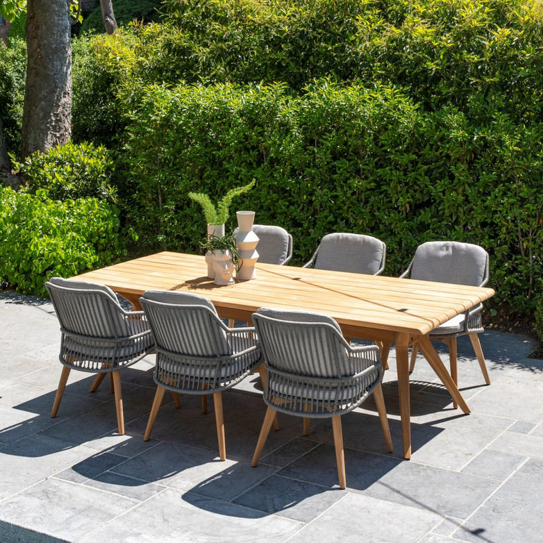 The Sempre 6 Seat Rectangular Dining Set with Teak Table in Light Grey is displayed on a patio, with six cushioned chairs, surrounded by lush green bushes.