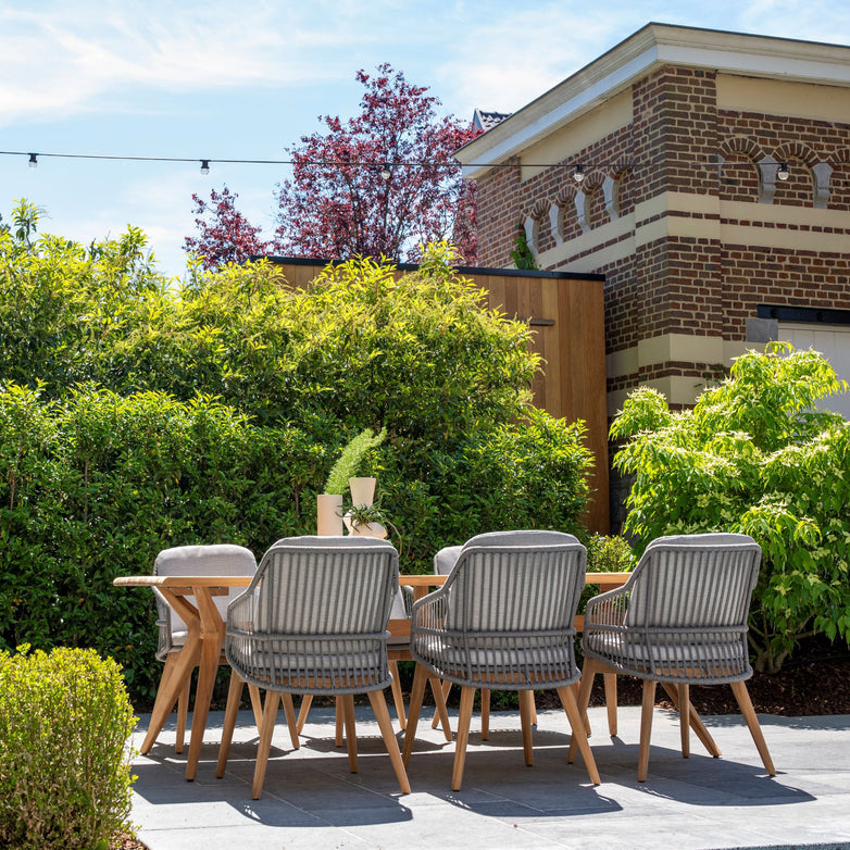 Sempre 6 Seat Rectangular Dining Set in Light Grey, featuring six grey chairs around a teak table, placed amidst green shrubs with a brick building in the background.