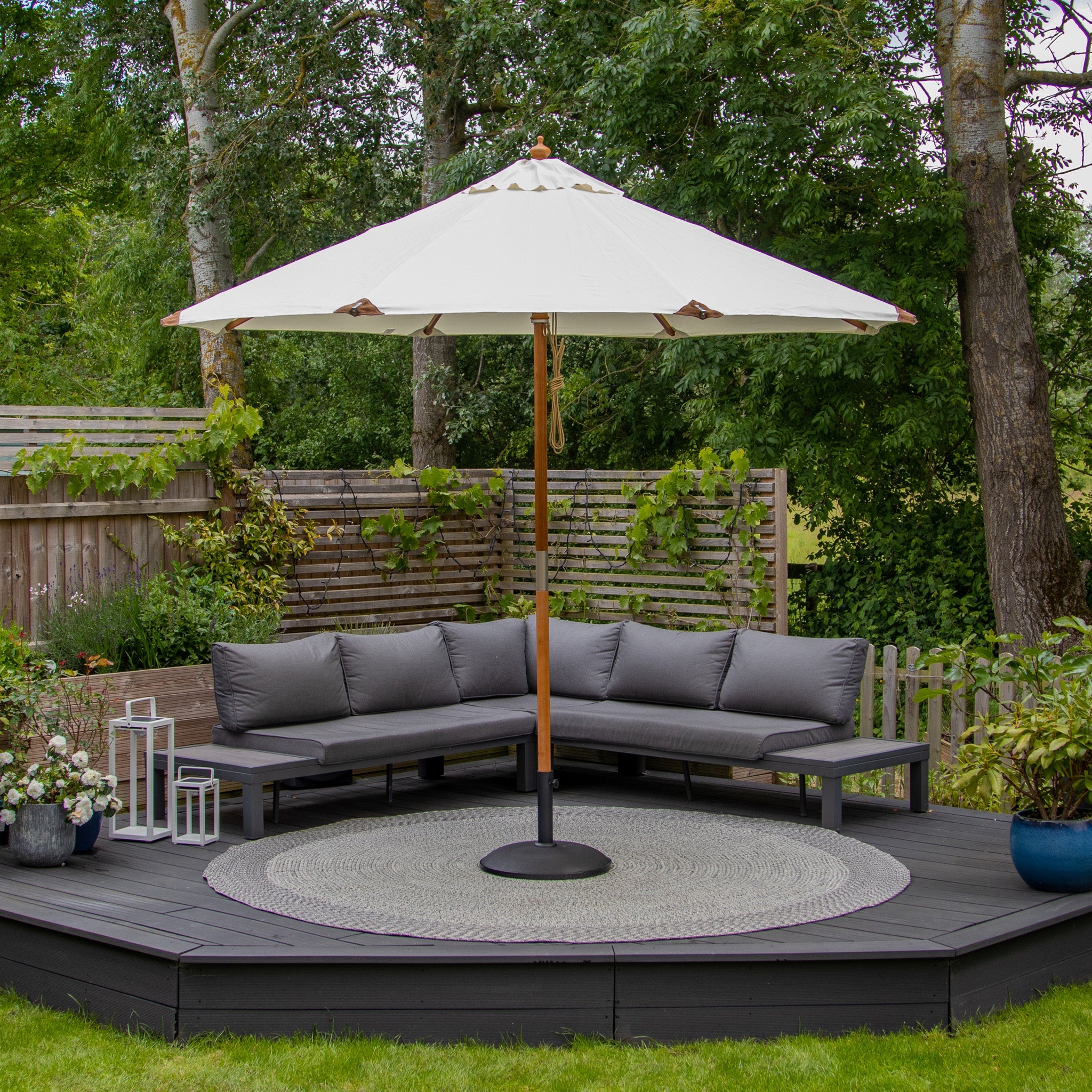 Outdoor seating area with a grey sectional sofa, a beige Styx 3m Round Deluxe Wooden Parasol, and a round rug on a wooden deck surrounded by greenery.