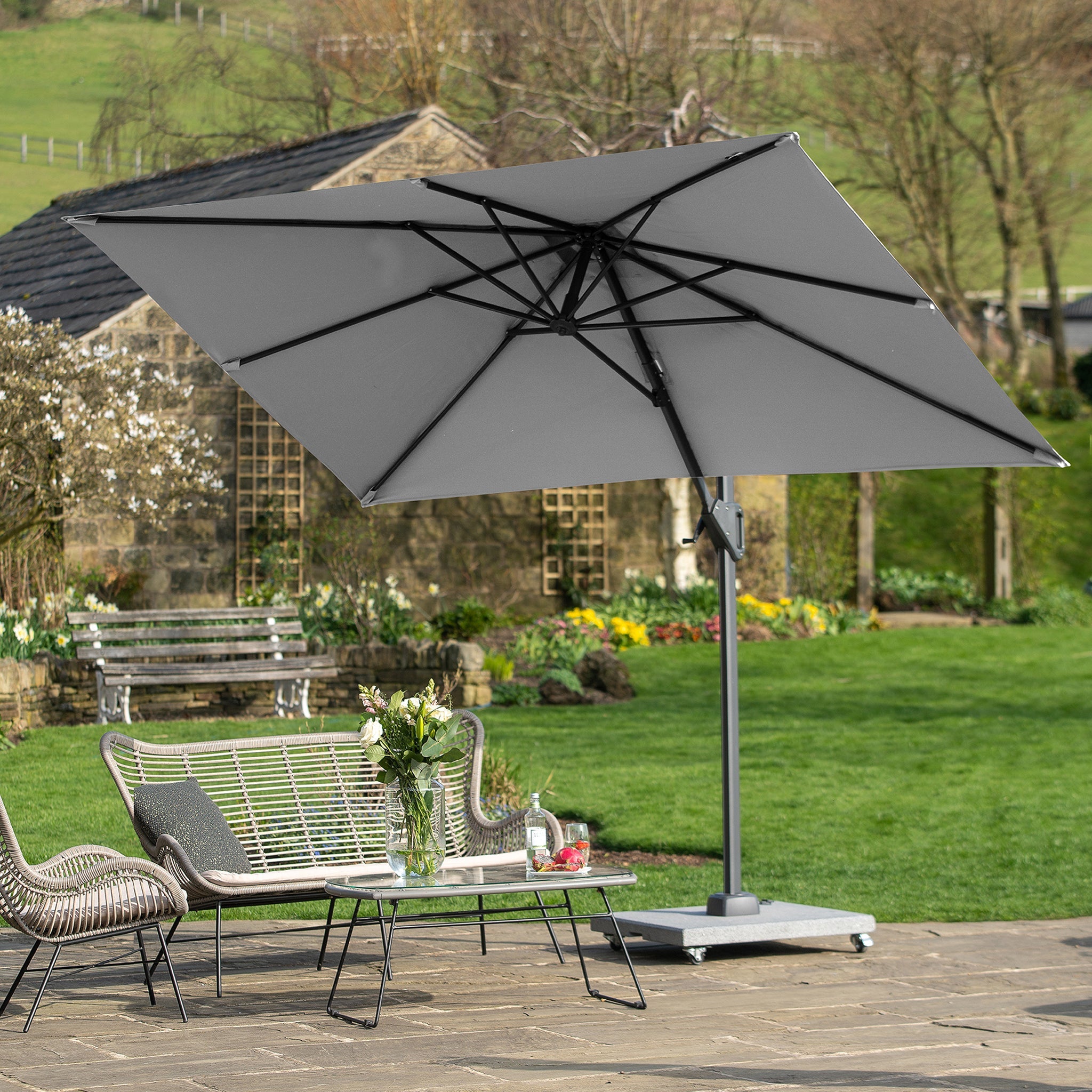 Outdoor seating area with metal benches and table under a large grey Voyager T1 3m x 2m Rectangular Parasol, featuring 360 degrees rotation, set on a stone patio in a garden.