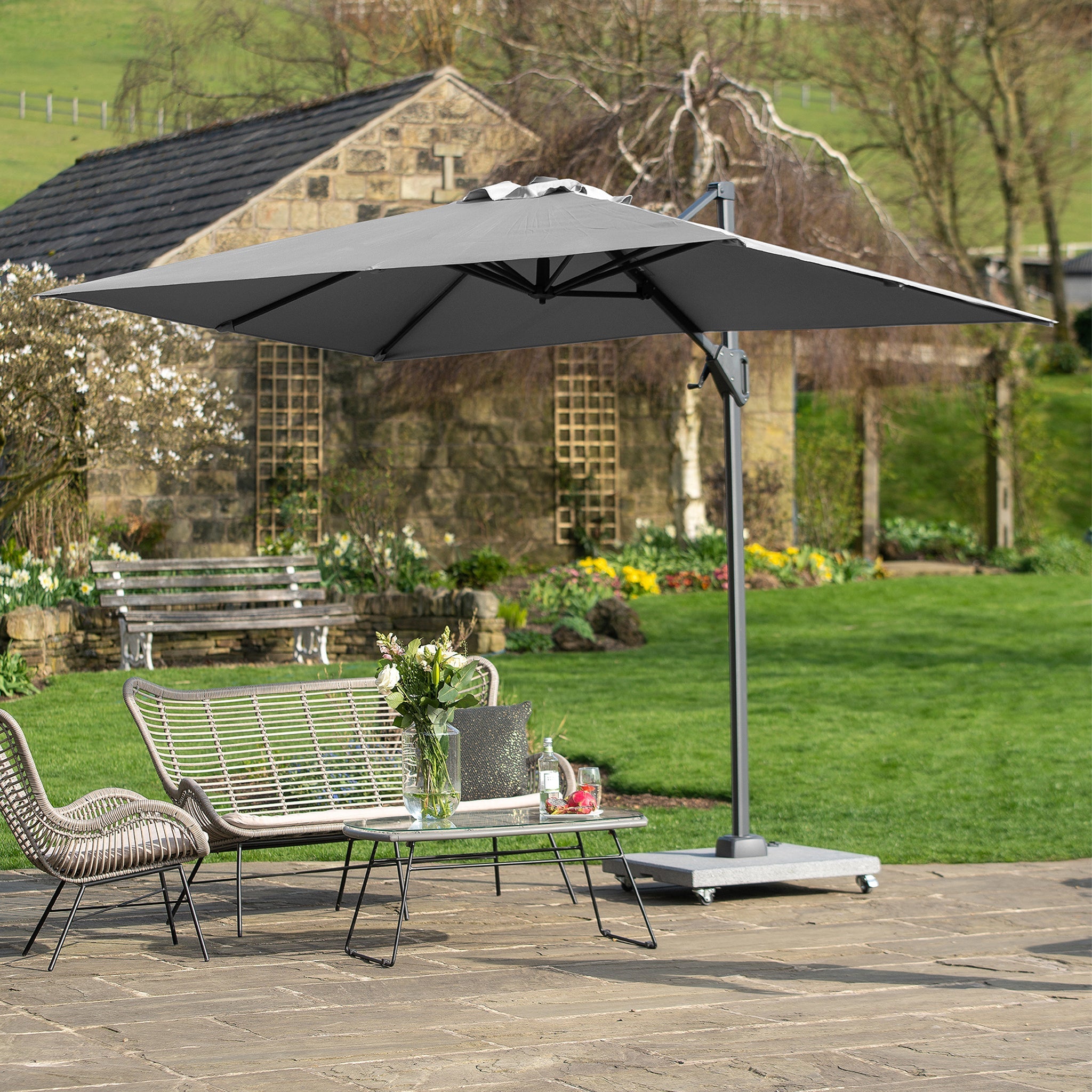 A garden patio featuring a table set, chairs, and a large gray Voyager T1 3m x 2m Rectangular Parasol that offers 360 degrees rotation. A rustic stone house and greenery can be seen in the background.