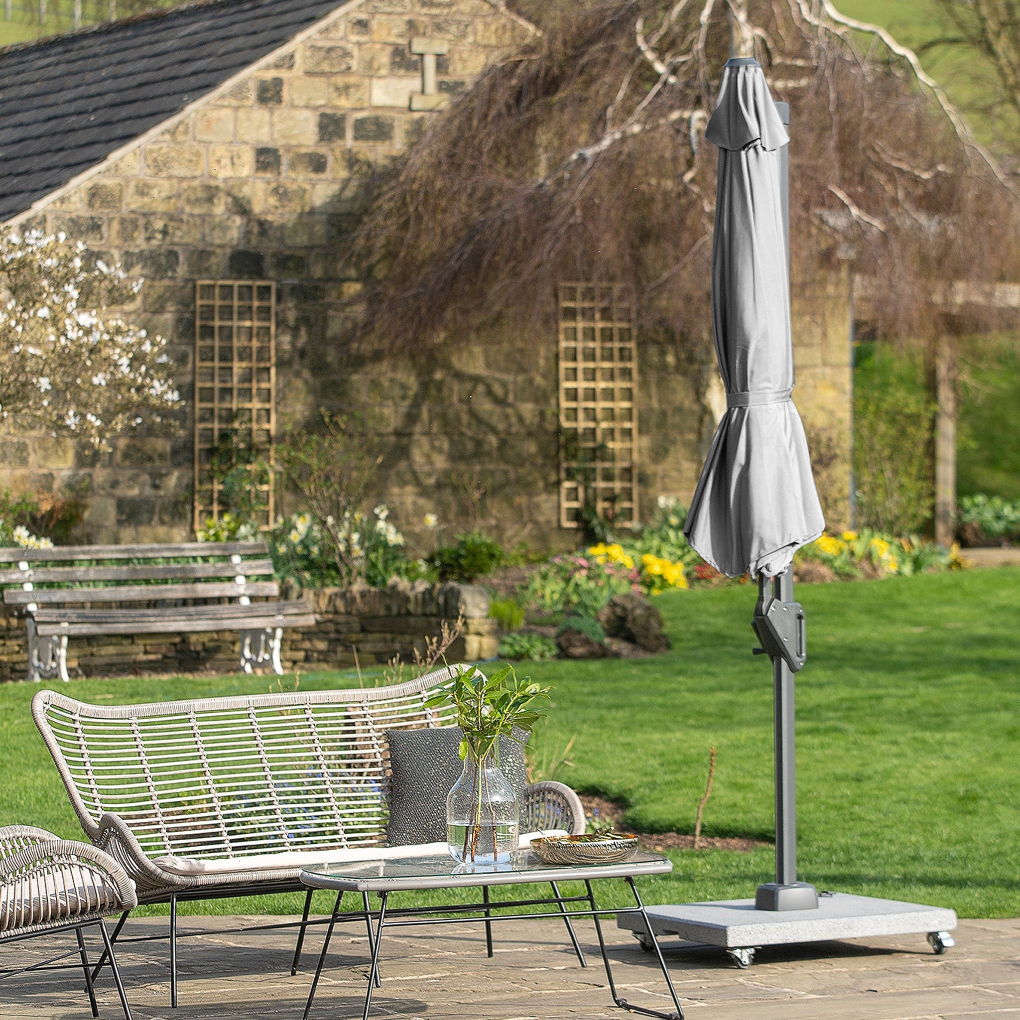 Patio garden with a woven bench and table near a closed Voyager T1 3m x 2m Rectangular Parasol in Grey. Stone building and trees in the background.