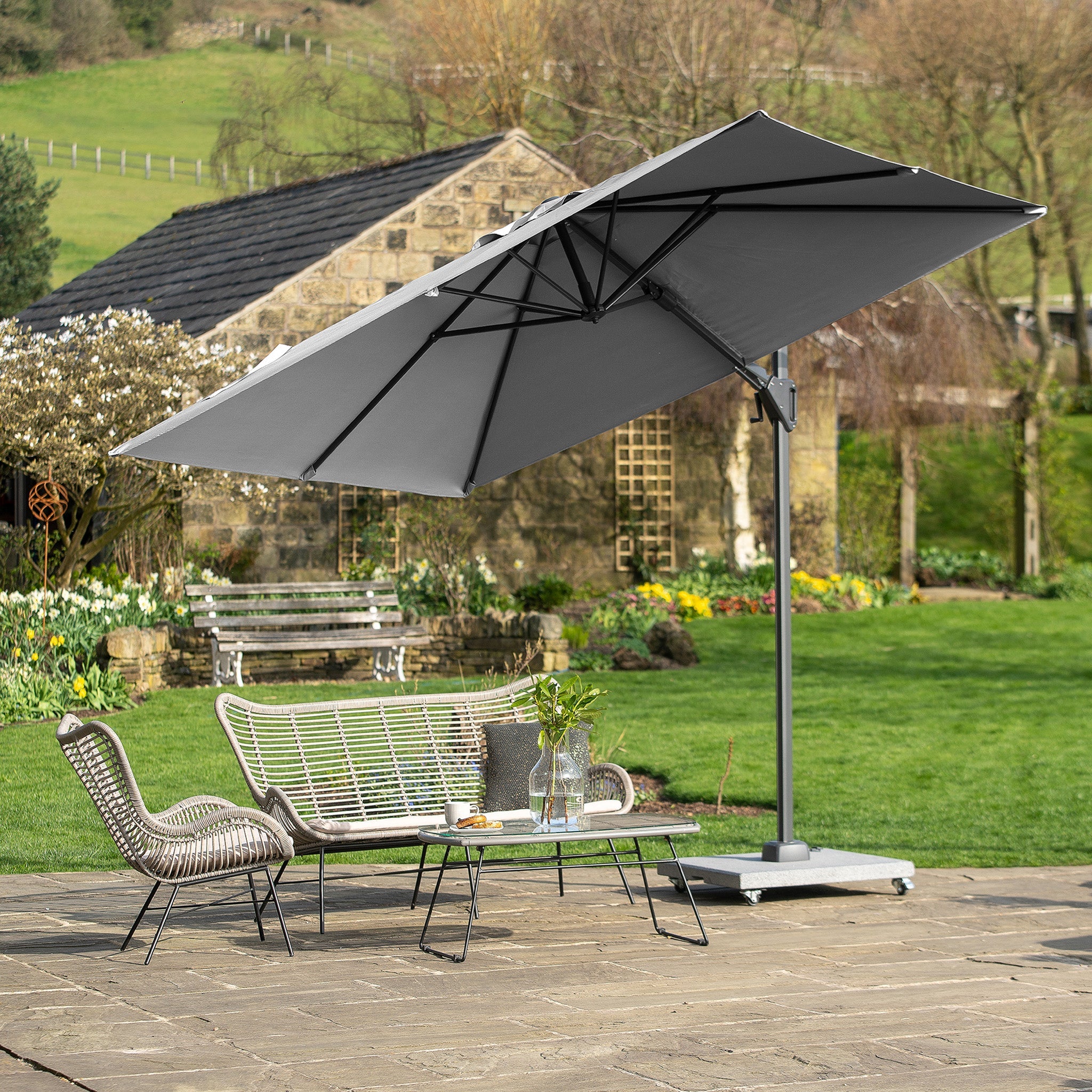 A patio with a Voyager T2 2.7m Square Parasol in Grey, featuring a 360° tilt and an aluminum pole, two chairs, and a coffee table, set in a lush garden with a stone building.