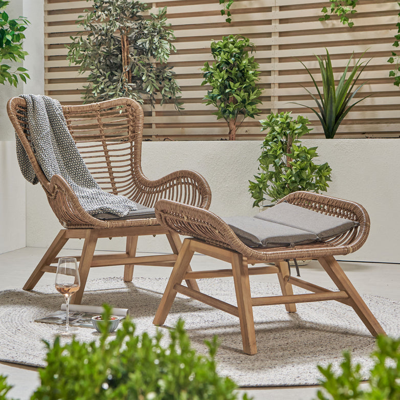 An Aurora Chair & Hocker Set Natural, showcasing a wicker chair adorned with a blanket and a matching ottoman on a round rug, sits next to a glass of wine in a lush outdoor setting, exuding retro glamour.