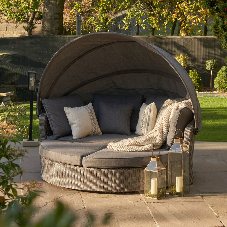 The Pacific Lifestyle Bermuda Daybed in Slate Grey has a round wicker design with cushions, a canopy, and two lanterns with candles. Perfect for patios, its weatherproof build ensures durability while creating a cozy retreat with garden views.