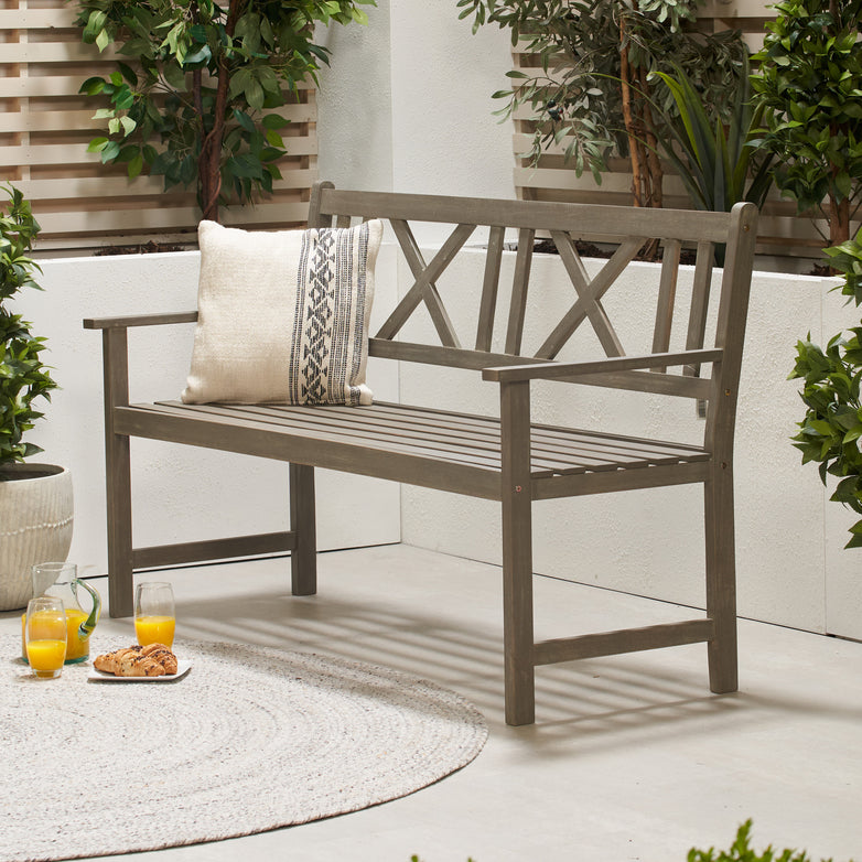 An Antique Grey Cambridge 3 Seater Acacia Wood Bench with a cushion in an outdoor patio area, complemented by garden furniture like a rug, orange juice, and snacks on the ground beside it.