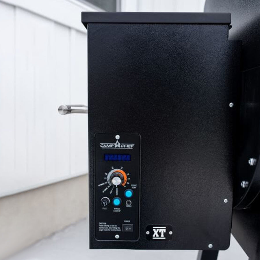 Close-up of a Camp Chef XT 24 Pellet Grill's digital control panel, featuring Smart Smoke Technology with buttons and knobs against a wall background.