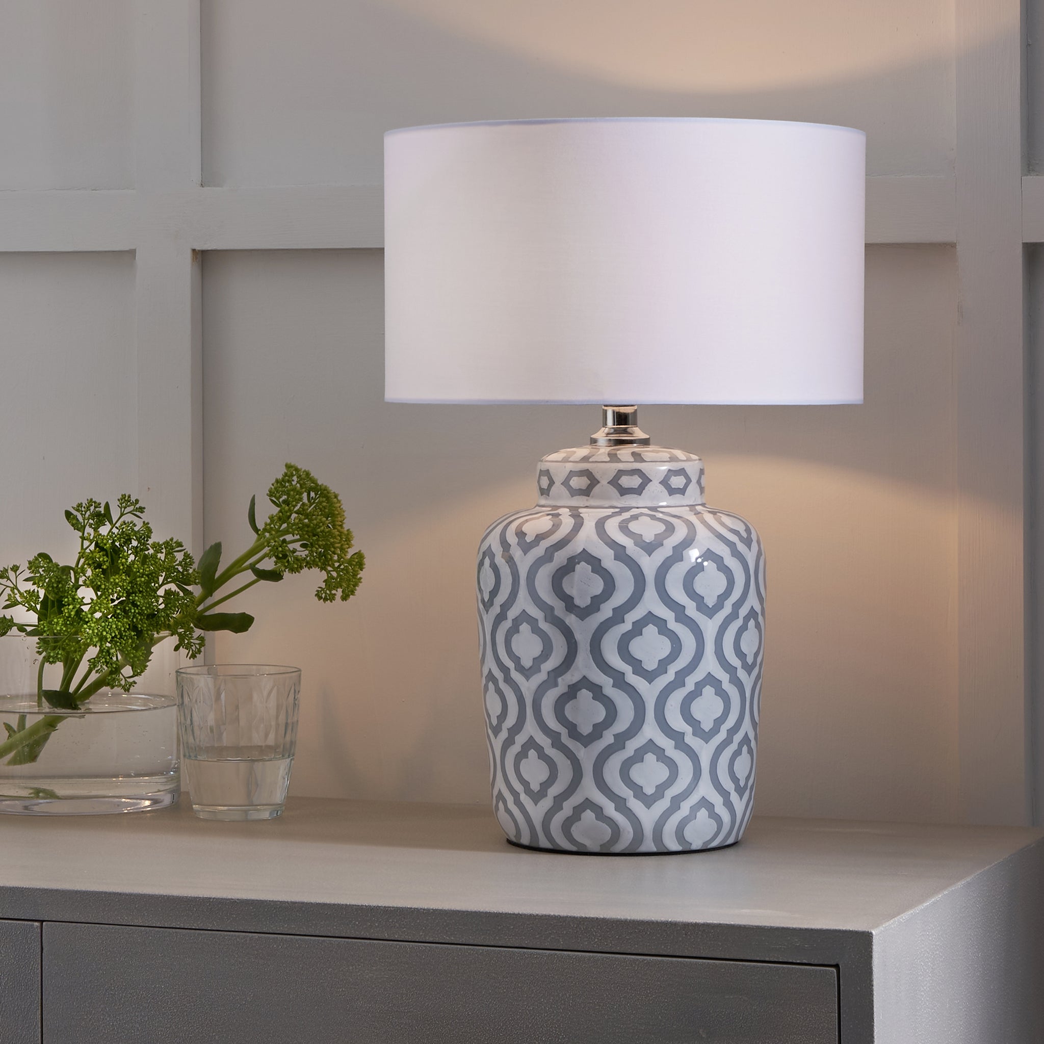 The Celia Grey and White Pattern Ceramic Table Lamp, with its classic shape and white shade, sits gracefully on a gray table next to a glass vase filled with green flowers.