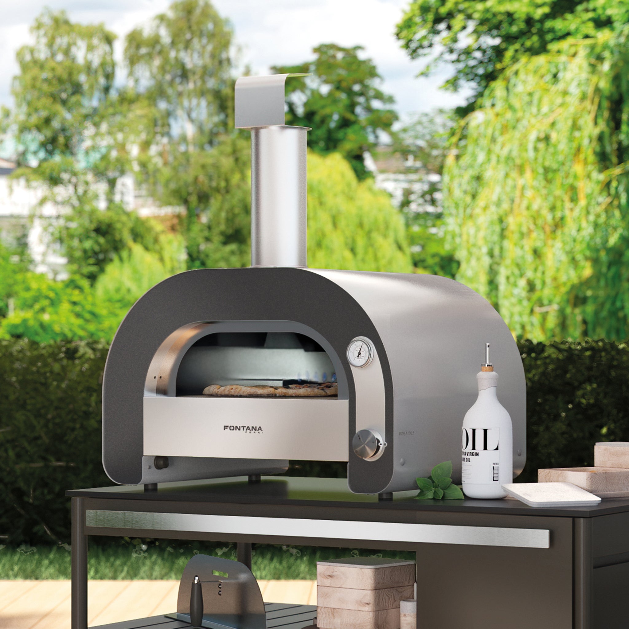 The Fontana Maestro 60 Gas Pizza Oven, set on a patio table surrounded by greenery, features an outdoor wood-fired design with a gas burner on the bottom.