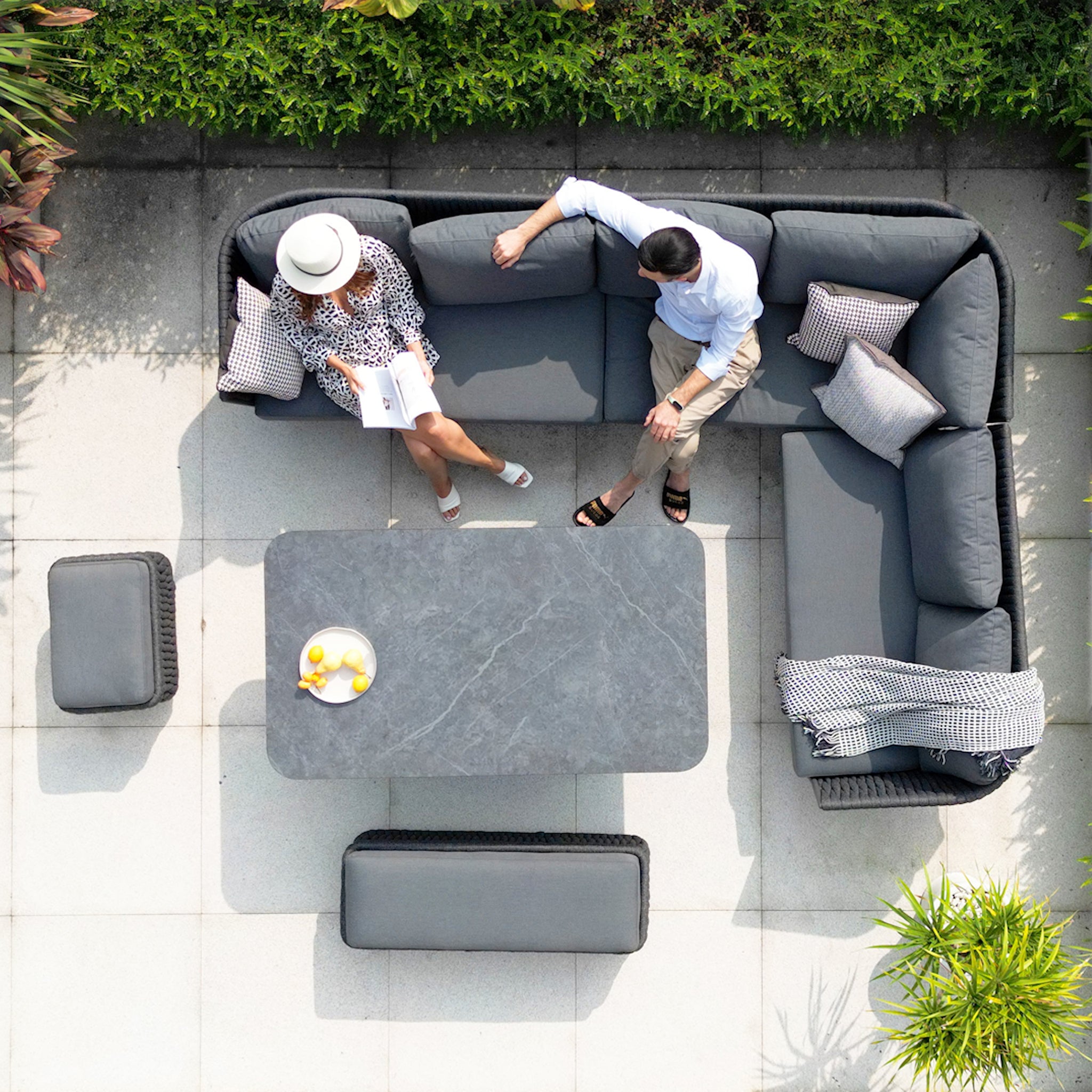 Top view of two people sitting on a gray Palma Rope Rectangular Corner Dining Set with Rising Table in Grey (Right Hand) with a table in the middle, surrounded by greenery. The sectional couch features a water-repelling thick rope weave and sturdy aluminum frames.