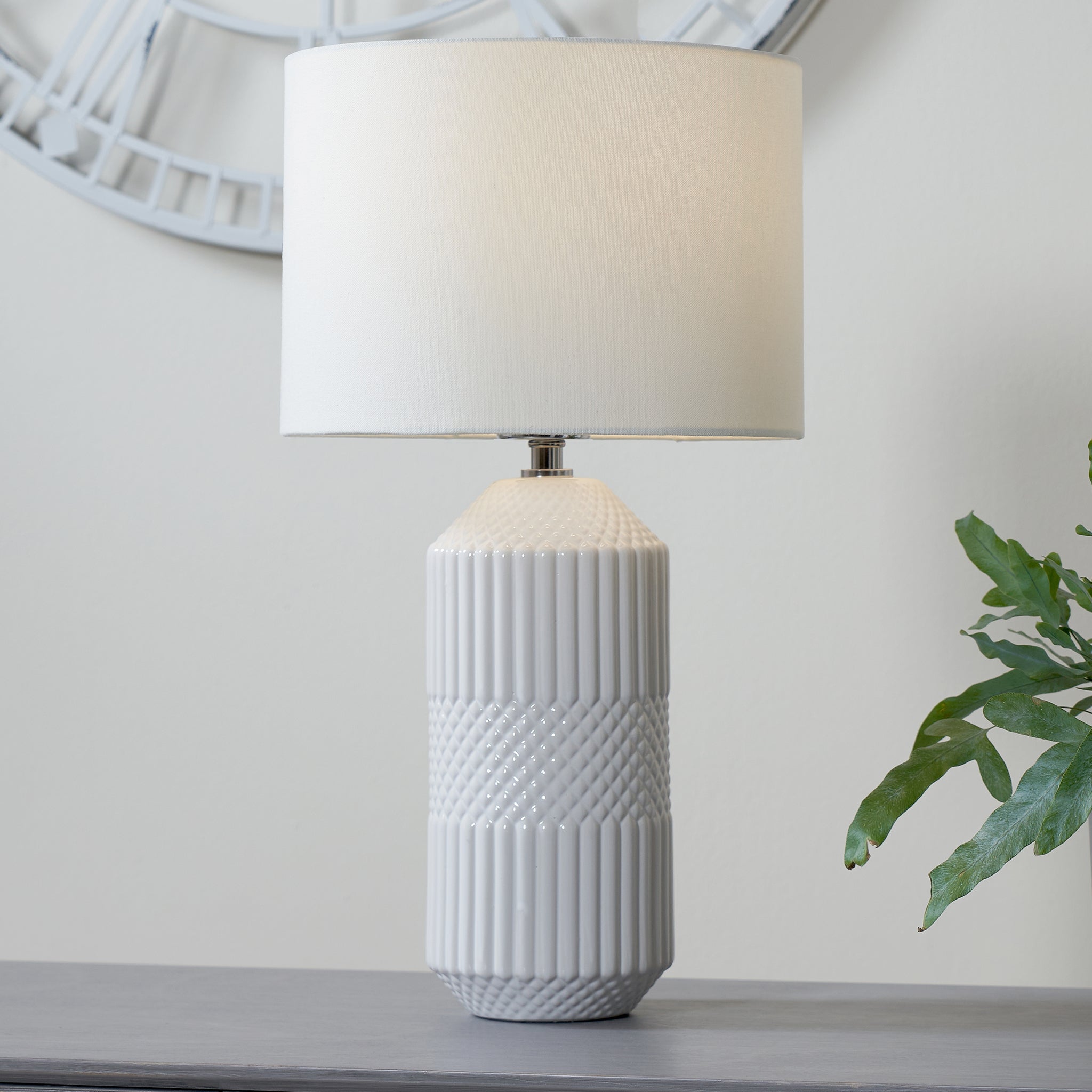 Meribel Geo Textured Tall Ceramic Table Lamp in White, featuring a white faux cotton cylindrical shade, placed on a grey surface with a green plant nearby.