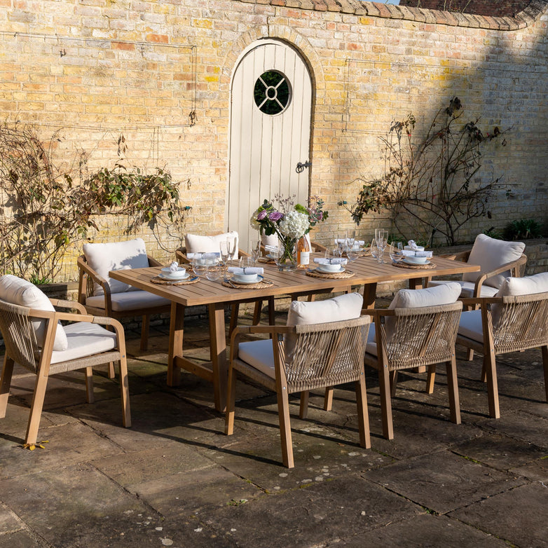 Introducing the Quay 8 Seat Rectangular Dining Set in Linen, featuring a beautifully crafted acacia wood table and eight chairs. Each piece is designed with water-resistant polyester fabric cushions for added comfort. Perfectly staged with elegant dishes and flowers, this outdoor dining set sits against a brick wall adorned with a charming wooden door.