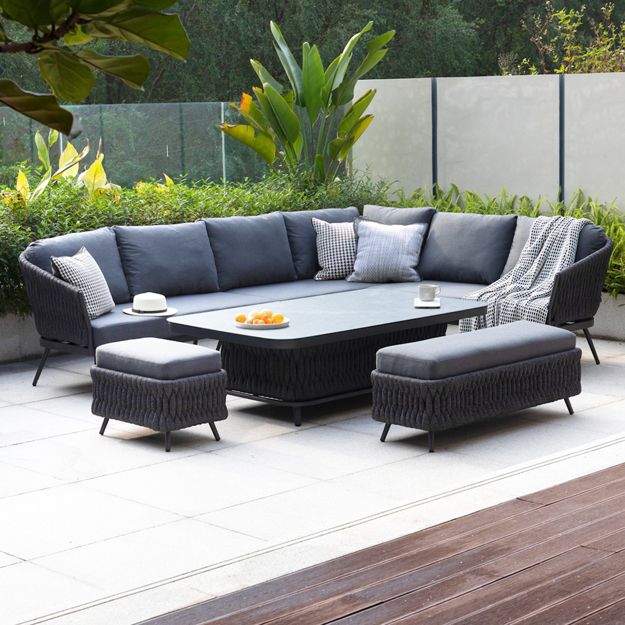 Outdoor patio featuring the Palma Rope Rectangular Corner Dining Set with Rising Table in Grey (Right Hand), showcasing a modern grey sectional sofa, coffee table, ottomans, and greenery in the background. The setup is complemented by aluminium frames and water-repelling thick rope weave for added durability and style.